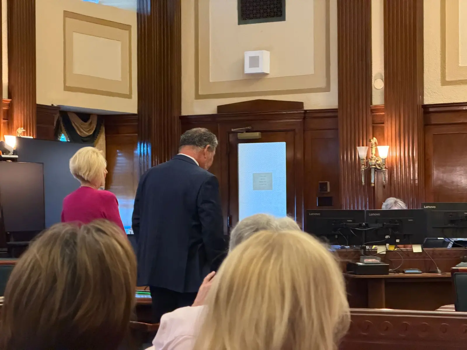 Brooke Covington, in a pink cardigan, stands with her attorney in a dark pinstripe suit facing the bench, seen from behind the courtroom pews.