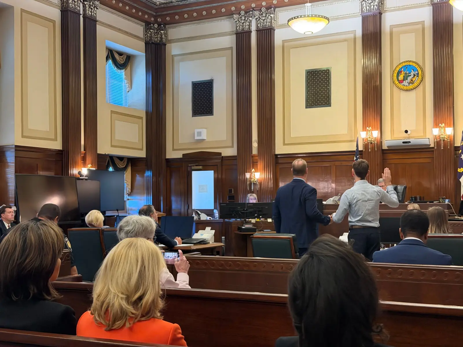 Matthew Fenner, in a light button-down shirt, raises his right hand to be sworn in before the judge, standing beside a man in a navy suit.