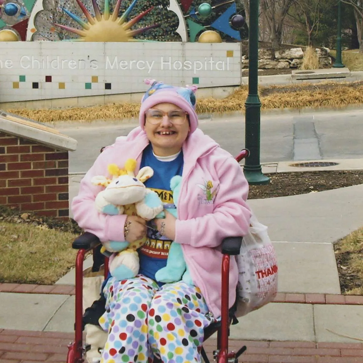 Gypsy Rose Blanchard smiling in a red wheelchair outside the Children's Mercy Hospital sign, wearing glasses, a pink knit hat, a pink fleece jacket and polka-dot pants, and holding stuffed animals.