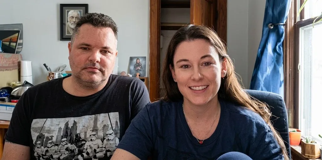Patrick Tomlinson, in a black T-shirt printed with Star Wars stormtroopers, sits beside a smiling Niki Robinson, in a navy T-shirt, in a home office with a window and blue curtains.